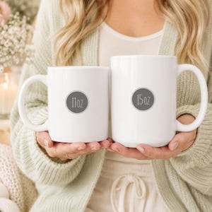 May include: Two white ceramic mugs with rounded handles. The smaller mug on the left features a grey circular graphic with the text 11oz, while the larger mug on the right features a grey circular graphic with the text 15oz printed in the center.