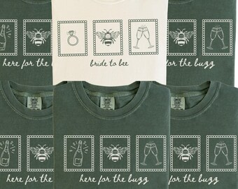May include: A collection of crew neck t-shirts in cream and dark green. The cream shirt features a ring, bee, and champagne glasses with the text bride to bee. The green shirts feature a champagne bottle, bee, and glasses with here for the buzz.
