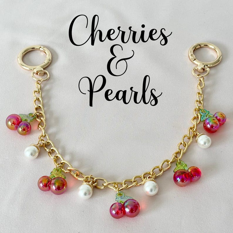May include: A gold chain keychain with red and green cherry charms and white pearl accents. The keychain has two gold rings at the ends. The text "Cherries & Pearls" is written above the keychain.