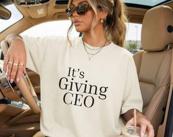 May include: Cream-colored oversized t-shirt with the text "It's Giving CEO" in black. The shirt is worn by a person in a car, accessorized with jewelry and sunglasses. The person is holding a beverage in a clear cup.