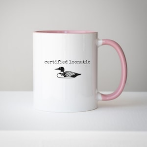 May include: A white ceramic mug with a light pink interior and handle. The exterior features a black graphic of a loon bird centered above the black typewriter-style text that reads certified loonatic in lowercase letters on the front side.