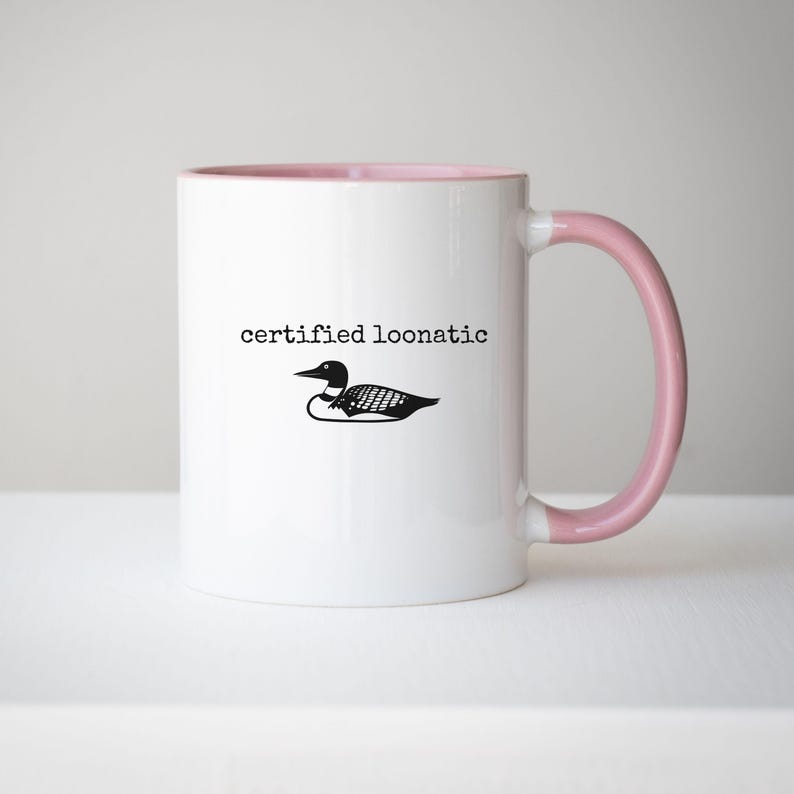 May include: A white ceramic mug with a light pink interior and handle. The exterior features a black graphic of a loon bird centered above the black typewriter-style text that reads certified loonatic in lowercase letters on the front side.