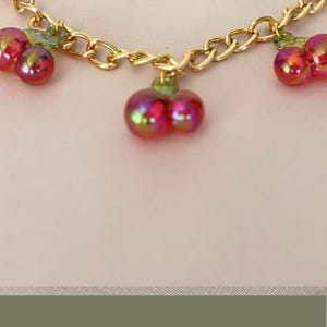 May include: A gold chain with red and green cherry charms. The charms are made of glass and have a shiny finish. The chain is perfect for adding a touch of whimsy to any bag.
