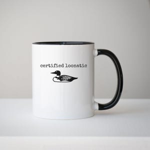 May include: A white ceramic mug with a black handle and interior rim. The front features a black graphic illustration of a loon bird positioned below the black text "certified loonatic" written in a typewriter-style font on the white surface.