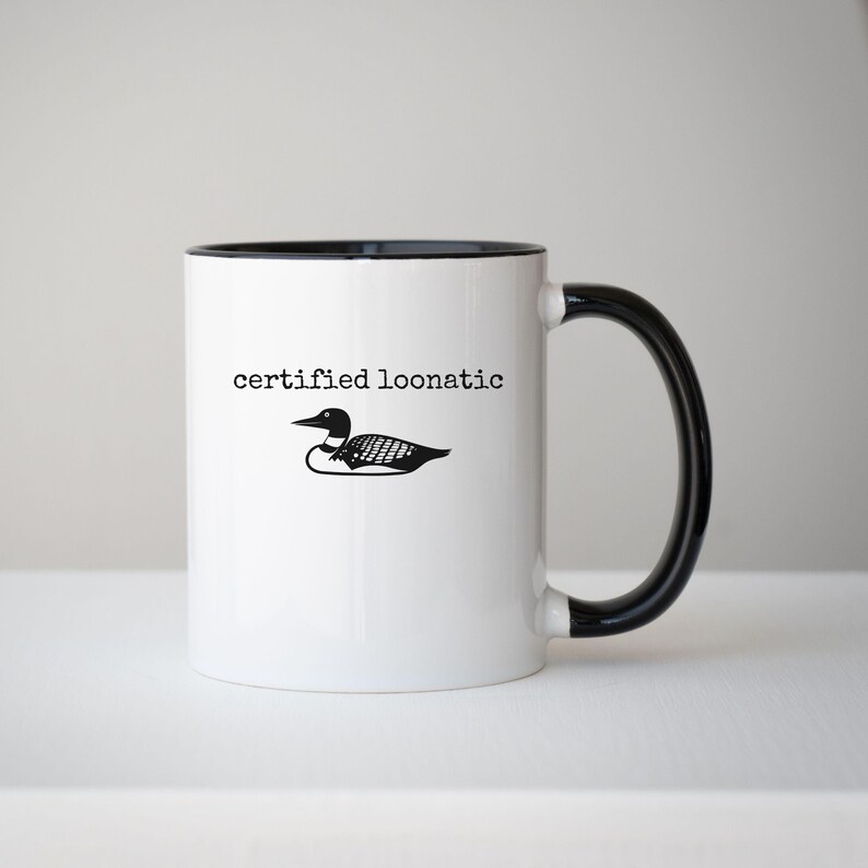May include: A white ceramic mug with a black handle and interior rim. The front features a black graphic illustration of a loon bird positioned below the black text "certified loonatic" written in a typewriter-style font on the white surface.