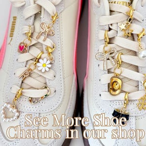 May include: Close-up of two white sneakers with colorful shoe charms attached to the laces. The charms include a heart, a daisy, a bow, a star, a cowboy hat, and a four-leaf clover. The text "See More Shoe Charms in our shop" is printed on the bottom of the image.