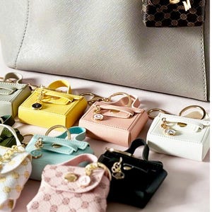 May include: A collection of miniature handbags in various colors and patterns, including yellow, pink, white, black, and green. The handbags are attached to keychains and have a small gold charm with a letter on it. The image is accompanied by the text "WHY ARE MINIATURES SO TRNEDY?"