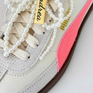 May include: A pair of white sneakers with pink accents and pearl necklaces tied around the laces. Each necklace has a gold tag with a name on it, "Andrea". The image is captioned "Shoe Charm Gifts".