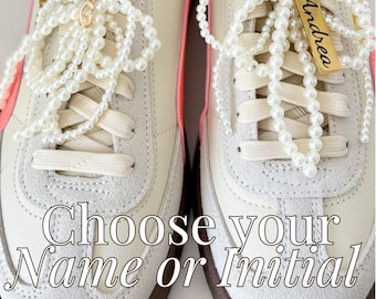 May include: A pair of white sneakers with pink accents and pearl laces. The laces are tied with a bow and a gold tag with the name "Andrea" is attached to the bow. The text "Choose your Name or Initial" is printed below the sneakers.