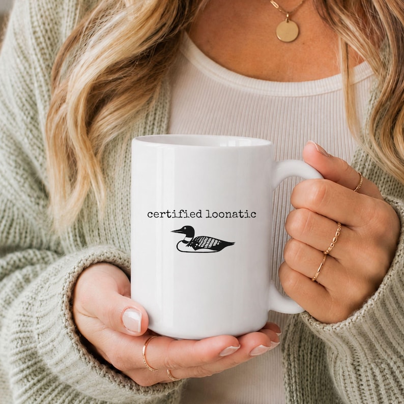May include: A white ceramic mug featuring a black graphic of a loon bird centered on the front. Above the bird, the text "certified loonatic" is printed in a simple, black, typewriter-style font. The mug has a smooth finish and a curved handle.