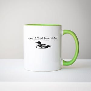 May include: A white ceramic mug with a bright green interior and handle. The exterior features a black graphic illustration of a loon bird centered above the black typewriter-style text that reads certified loonatic in lowercase letters.