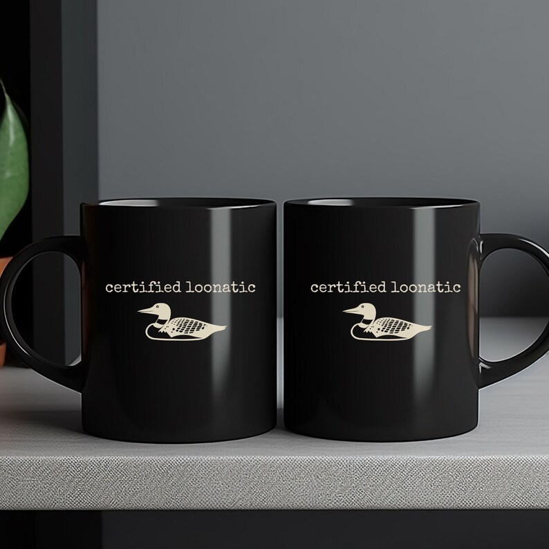 May include: Two black ceramic mugs featuring the text "certified loonatic" in a typewriter-style font above a graphic illustration of a loon bird in cream color. The mugs have a smooth, glossy finish and a standard handle on the side.