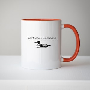 May include: A white ceramic mug with an orange interior and handle. The front features a black graphic of a loon bird positioned below the black text "certified loonatic" written in a typewriter-style font. The mug has a smooth, glossy finish.