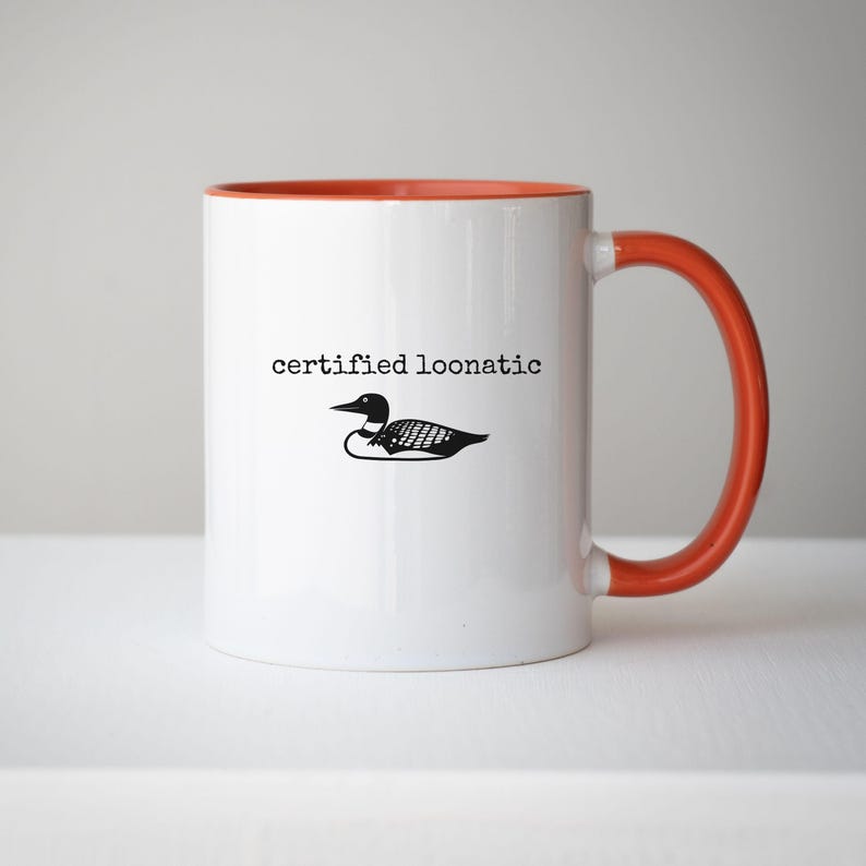 May include: A white ceramic mug with an orange interior and handle. The front features a black graphic of a loon bird positioned below the black text "certified loonatic" written in a typewriter-style font. The mug has a smooth, glossy finish.