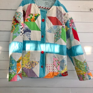 May include: A colorful patchwork jacket with a variety of fabric patterns, including floral, polka dot, and gingham. The jacket features a light blue trim and red buttons. The jacket is hanging on a white wooden wall.
