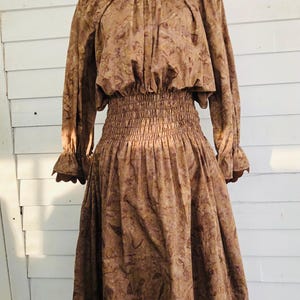 May include: A brown dress with a floral pattern. The dress has long sleeves with ruffled cuffs, a gathered neckline, and a smocked waist. The skirt is full and falls to the mid-calf. The dress is displayed on a black mannequin.
