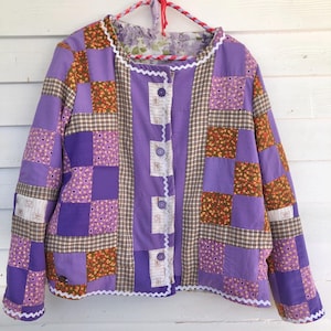 May include: A patchwork jacket in shades of purple, brown, and floral patterns. The jacket has a button-up front, white scalloped trim, and a floral lining. It is hanging on a red and white rope hanger.