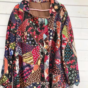 May include: A patchwork jacket with a button-up front, hanging on a pink hanger. The jacket is made from various patterned fabrics in red, blue, brown, and green. The collar and lining are a solid brown colour.