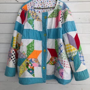 May include: A patchwork jacket with a button-up front, displaying a vibrant mix of patterns and colours. The jacket incorporates floral, polka dot, and geometric designs in red, orange, blue, green, and white. It is suspended from a wooden hanger.