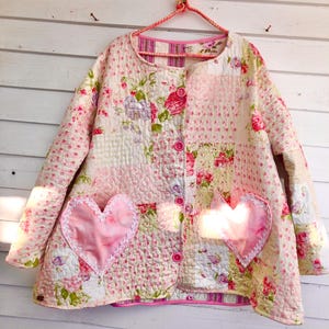 Puede incluir: Chaqueta acolchada con diseño patchwork con estampados florales en rosa, blanco y verde. La chaqueta tiene dos bolsillos en forma de corazón con ribete de encaje y botones rosas. Cuelga de una percha rosa.