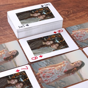 Op de afbeelding: Een pak speelkaarten met aangepaste foto's. De kaarten hebben een witte rand en standaard symbooltekens. De afbeeldingen op de kaarten tonen twee personen. De kaarten zijn gerangschikt op een houten oppervlak.