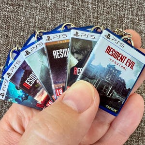 Può includere: Una collezione di portachiavi rettangolari in miniatura progettati per assomigliare a custodie di giochi per PlayStation 5. Ogni portachiavi è dotato di un piccolo anello metallico e mostra la copertina di titoli Resident Evil, incluso Resident Evil Requiem, con il logo PS5.