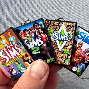Peut inclure: Quatre porte-clés représentant les couvertures des jeux The Sims : The Sims, The Sims 2, The Sims 3 et The Sims 4. Chaque porte-clés a un anneau argenté et présente les illustrations colorées du jeu, avec des personnages et des logos. Les porte-clés sont tenus dans une main.