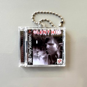 Puede incluir: Un llavero de plástico transparente con una réplica en miniatura de la caja del juego "Silent Hill" de PlayStation. La caja del juego tiene una imagen en blanco y negro de la cara de un hombre y el título del juego. El llavero tiene una cadena de bolas plateada.