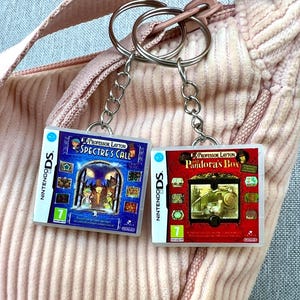 May include: Two keychains featuring miniature game cases for Nintendo DS games. One keychain has a blue case with the title "Professor Layton and the Spectre's Call." The other keychain has a red case with the title "Professor Layton and Pandora's Box."