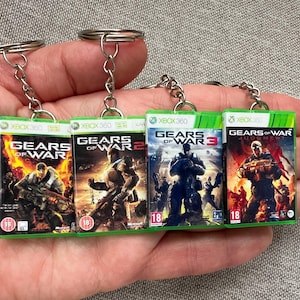 Puede incluir: Cuatro llaveros con miniaturas de carátulas de juegos de la serie "Gears of War" para Xbox 360. Cada llavero muestra una portada de juego diferente, con diseños verdes y negros vibrantes. Los llaveros están unidos a anillos metálicos plateados.
