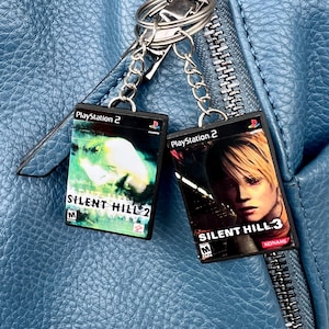 Puede incluir: Dos llaveros con carcasas de juegos de PlayStation 2 en miniatura. Uno muestra la carátula de "Silent Hill 2", el otro de "Silent Hill 3". Ambos llaveros están sujetos a un llavero plateado.