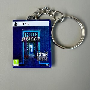 以下が含まれることがあります： 「Blue Prince: Day One Edition」のPS5ゲームケースデザインの青いキーホルダー。キーホルダーはシルバーのチェーンとキーリングが付いています。ゲームケースのアートワークは、ドアの中にいる人物を描いています。