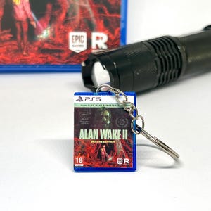 Könnte beinhalten: Ein blauer und weißer Videospiel-Schlüsselanhänger mit dem Cover von Alan Wake II: Deluxe Edition. Der Schlüsselanhänger enthält eine silberne Kette und einen silbernen Ring.
