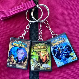 Könnte beinhalten: Drei World of Warcraft DVD-Hüllen-Schlüsselanhänger. Jeder Schlüsselanhänger zeigt ein anderes Spielcover mit lebendigen Grafiken und dem Spieletitel. Die Schlüsselanhänger sind an silbernen Schlüsselringen befestigt und hängen vor einem rosa Hintergrund.