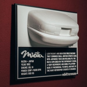 Peut inclure: Un modèle 3D imprimé en blanc d'une Mazda Miata, une voiture de sport légère, monté sur une plaque noire avec des détails sur la voiture, y compris l'année, le moteur, la puissance et le poids. La plaque comprend également le texte "Miata" dans une police stylisée.