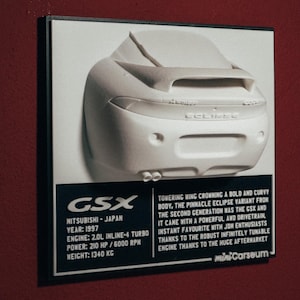 Puede incluir: Un modelo blanco de un coche Mitsubishi Eclipse GSX de 1997 con un gran alerón trasero. El modelo se muestra sobre un fondo negro con texto que dice "GSX", "Mitsubishi - Japón", "Año: 1997", "Motor: 2.0L Inline-4 Turbo", "Potencia: 210 CV / 6000 RPM", "Peso: 1340 KG", "Ala imponente coronando una carrocería audaz y curvada, la variante Eclipse por excelencia de la segunda generación fue la GSX y venía con un tren motriz potente y eficaz. Un favorito instantáneo entre los entusiastas de Jon gracias al motor robusto infinitamente sintonizable gracias al enorme mercado de accesorios... miniCarseum".