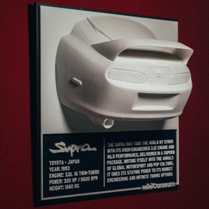 以下が含まれることがあります： 銀色のフォントで「Supra」と書かれた黒いプレートに、1993年製の白いトヨタスープラのモデルカーが取り付けられています。プレートには、車の仕様（年式、エンジン、出力、重量）も記載されています。プレートには、「THE SUPRA NA TOOK THE WORLD BY STORM WITH ITS OVER-ENGINEERED 2JZ ENGINE AND WILD PERFORMANCE, DELIVERED IN A SUPERB PACKAGE, WRITING ITSELF INTO THE ANNALS OF GLOBAL MOTORSPORT AND POP CULTURE, IT OWES ITS STAYING POWER TO ITS ROBUST ENGINEERING AND INFINITE TUNING OPTIONS」というテキストが印刷されています。