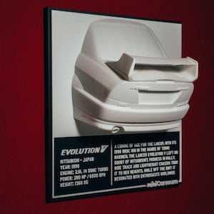 Puede incluir: Modelo 3D blanco de la parte trasera de un coche, montado en un marco negro. El modelo tiene un alerón y está sobre un fondo blanco. El texto en una etiqueta negra incluye "EVOLUTION V" y las especificaciones del coche.