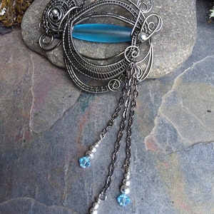 May include: A silver necklace with a blue glass pendant and dangling crystal beads. The pendant is made of wire wrapped in a circular design.