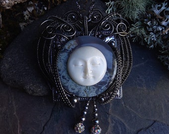 May include: A silver wire wrapped pendant with a carved moon face cameo set in a dark gray agate stone. The pendant hangs from a silver chain with two crystal beads.