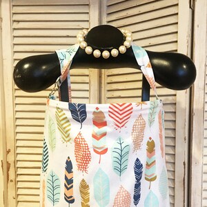 May include: A white bib with a colorful feather pattern. The bib has adjustable straps and is made of a soft, comfortable fabric.