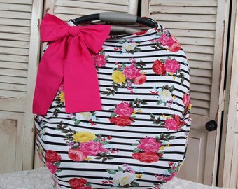 Puede incluir: Una funda para asiento de coche de bebé con un patrón de rayas blancas y negras, adornada con diseños florales rosas, rojos y amarillos. Un gran lazo rosa brillante está sujeto en la parte superior. La funda está diseñada para un asiento de coche.