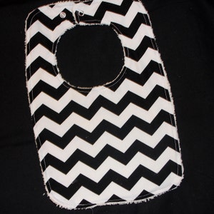 May include: A baby bib with a black and white chevron pattern. The bib has a rounded neck opening and two snap closures at the top. The edges are finished with a soft, white trim.