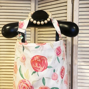 May include: A white apron with a pink and red floral pattern. The apron has adjustable straps and is hanging on a black hanger. A pearl necklace is on the hanger.