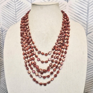 May include: A multi-strand necklace with reddish-brown beads. The necklace is displayed on a mannequin. The beads are small, flat, and have a slightly iridescent quality. The necklace has six strands of varying lengths.