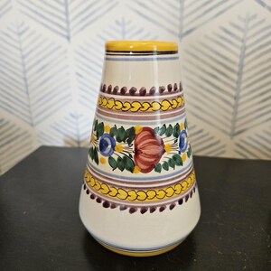 Puede incluir: Un jarrón de cerámica blanca con borde y base amarillos, adornado con motivos florales y geométricos en rojo, azul y amarillo. El jarrón tiene una forma ligeramente acampanada, adecuado para exhibir flores o como pieza decorativa.