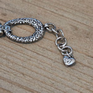 K&ouml;nnte beinhalten: Nahaufnahme eines silberfarbenen Armbandverschlusses und einer Kette. Der Verschluss hat eine ovale Form mit einem verzierten, wirbelnden Design. Die Kette hat kleine, runde Glieder und einen herzf&ouml;rmigen Anh&auml;nger.