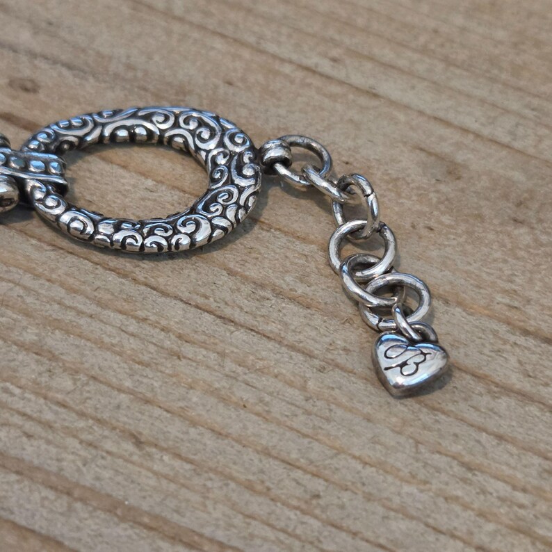 K&ouml;nnte beinhalten: Nahaufnahme eines silberfarbenen Armbandverschlusses und einer Kette. Der Verschluss hat eine ovale Form mit einem verzierten, wirbelnden Design. Die Kette hat kleine, runde Glieder und einen herzf&ouml;rmigen Anh&auml;nger.