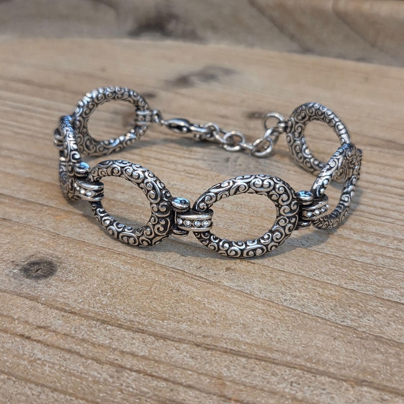 K&ouml;nnte beinhalten: Silberfarbenes Armband mit ovalen Gliedern, verziert mit filigranen Verzierungen und kleinen, klaren Edelsteinen. Das Armband hat einen Karabinerverschluss. Das Schmuckst&uuml;ck liegt auf einer Holzoberfl&auml;che.