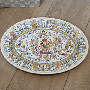 Könnte beinhalten: Ovale Servierplatte aus Keramik mit handbemaltem Design. Die Mitte zeigt einen Hahn, umgeben von floralen und scrollartigen Elementen in Gelb, Blau und Orange. Der Rand hat eine dekorative Bordüre. Ein dekoratives Stück zum Servieren von Speisen.